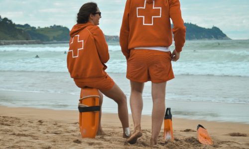 Two beach lifeguards in orange uniforms standing on the sand beside a rescue board and fins