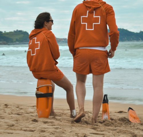 Two beach lifeguards in orange uniforms standing on the sand beside a rescue board and fins