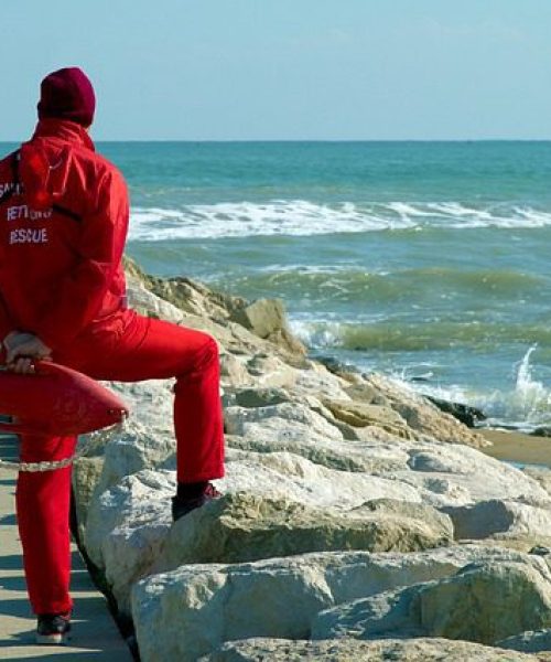 Lifeguard in a red uniform sitting on a rock breakwater with a rescue tube, watching the sea