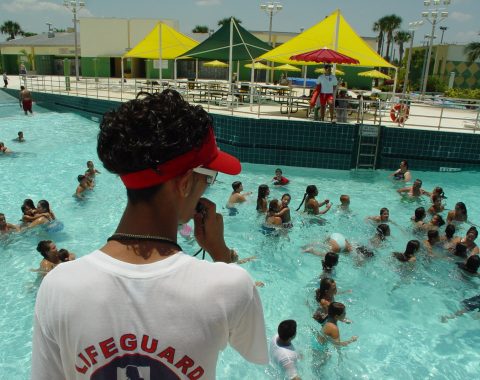 Lifeguard in a white lifeguard shirt and red visor watching swimmers in a busy outdoor pool