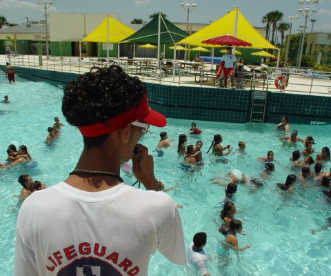Lifeguard in a white lifeguard shirt and red visor watching swimmers in a busy outdoor pool