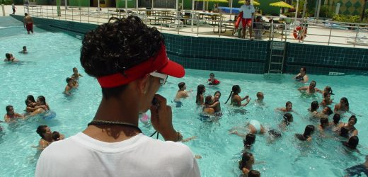 Lifeguard in a white lifeguard shirt and red visor watching swimmers in a busy outdoor pool